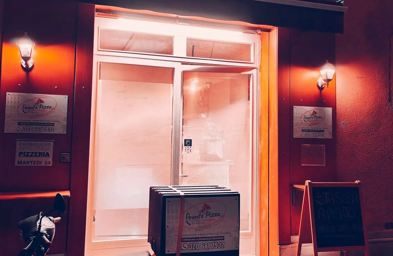 La vetrina della pizzeria illuminata di sera, con lo scooter delle consegne davanti